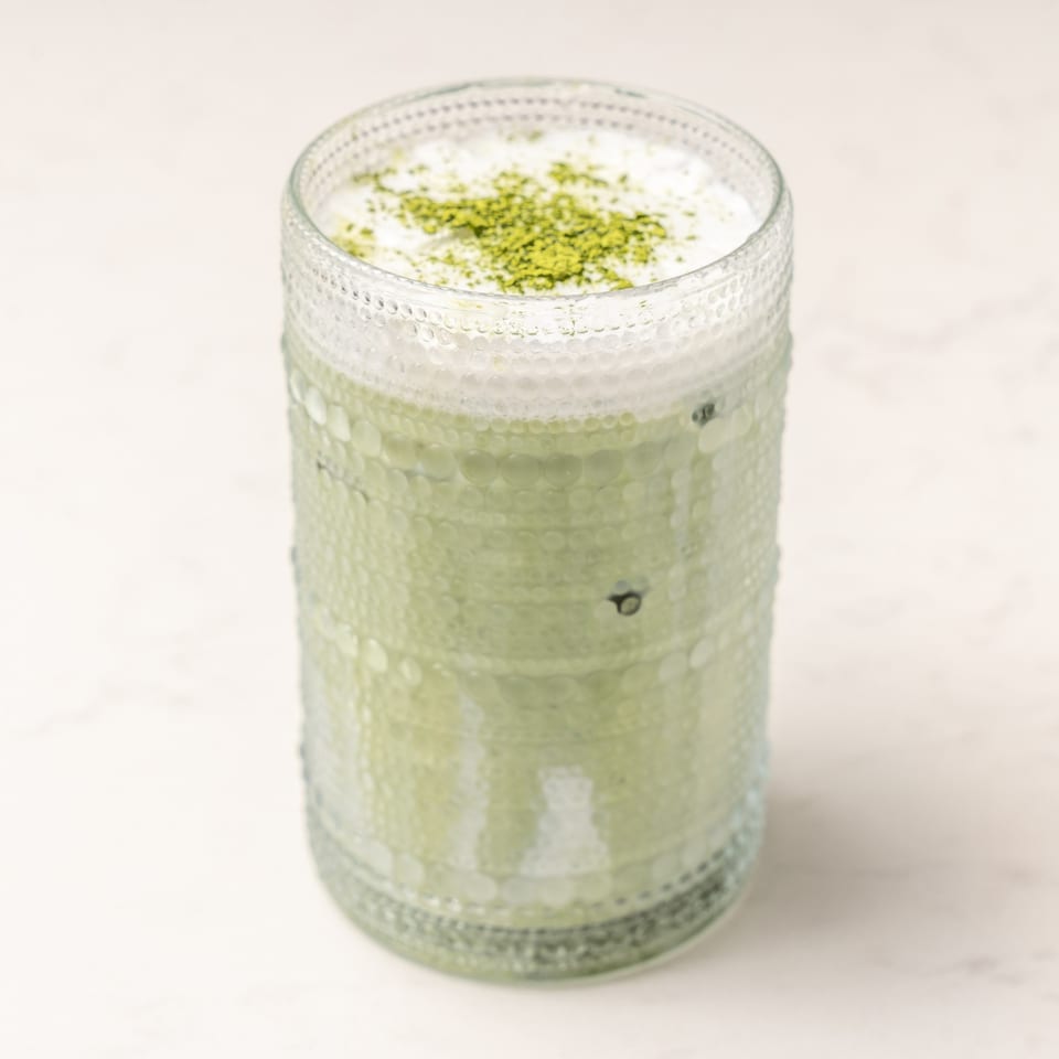 A tall glass of green matcha latte with foam and matcha powder on top.