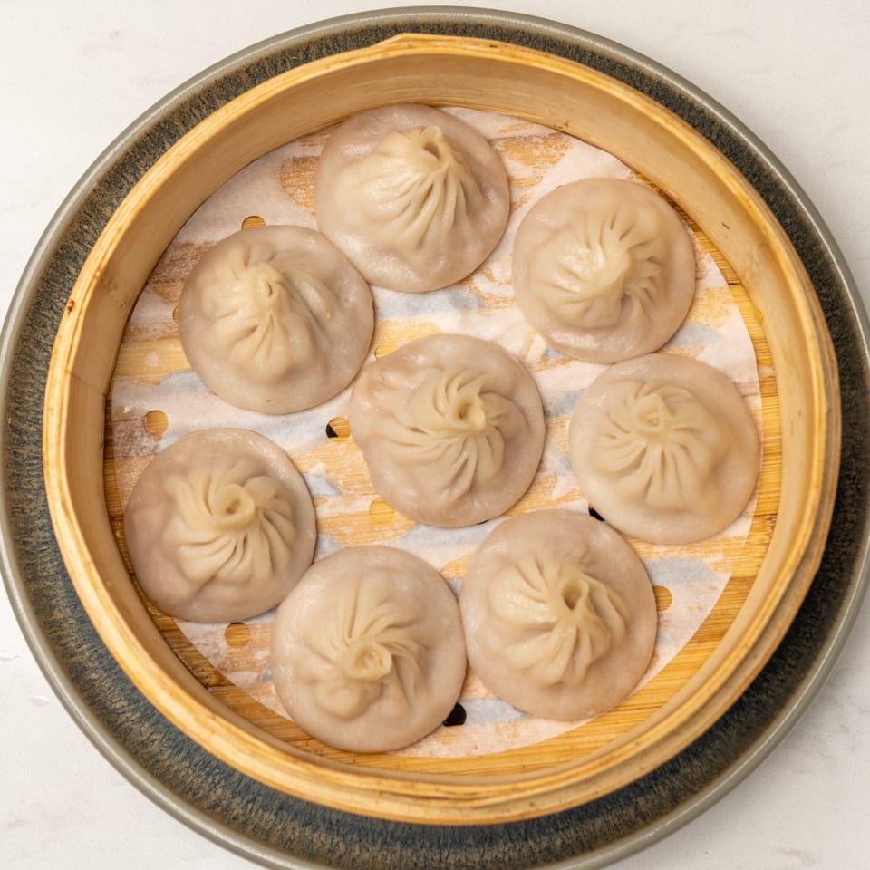 Eight steamed dumplings, likely Xiao Long Bao, arranged in a bamboo steamer basket.