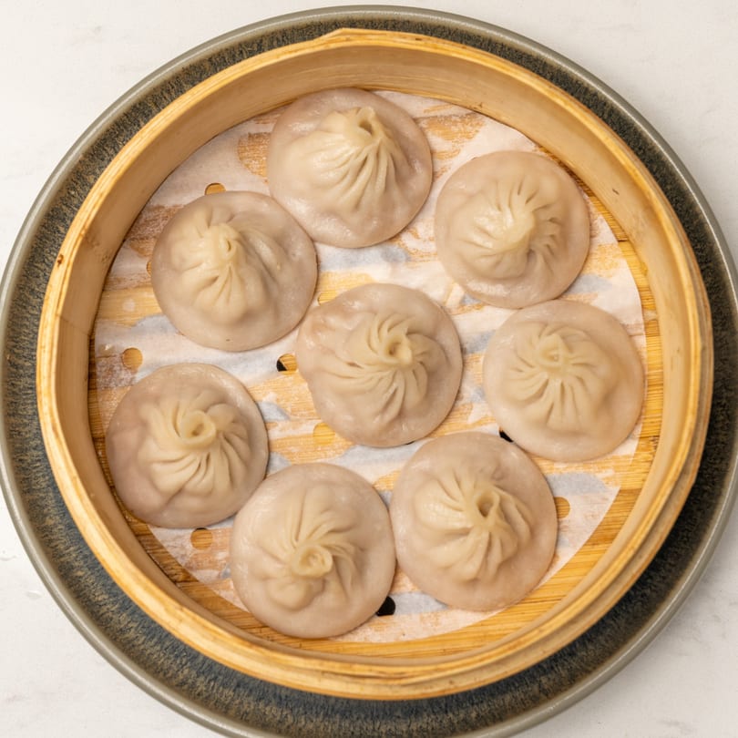 Eight steamed dumplings, likely Xiao Long Bao, arranged in a bamboo steamer basket.