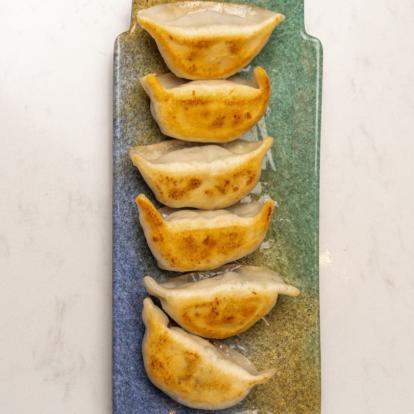 Six pan-fried dumplings with golden-brown bottoms, arranged on a long, rectangular, blue and green speckled plate.