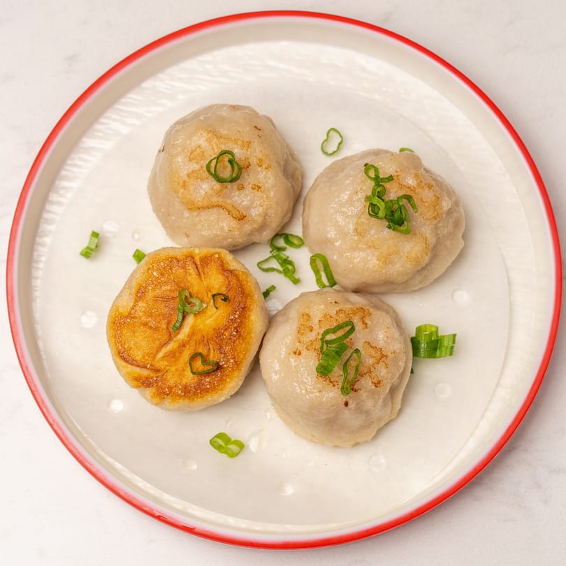Four pan-fried dumplings, one golden brown, garnished with green onions on a white plate with a red rim.